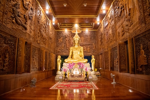 A large, golden Buddha statue sits at the center of an ornately decorated hall. The walls are adorned with intricate wooden carvings depicting various scenes and figures. Two smaller statues flank the central Buddha, and a variety of offerings and flowers are placed in front of it on a red and gold rug. The ceiling is wooden with bright lights illuminating the entire area.