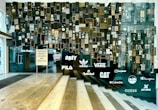A wall art installation composed of numerous vintage speakers of various sizes and designs, interspersed with light-up brand logos such as Adidas, Vans, and Timberland, among others. The display includes a sign listing product categories like womenswear, menswear, and accessories. The setting appears to be a modern retail environment, featuring reflective surfaces and glass elements.