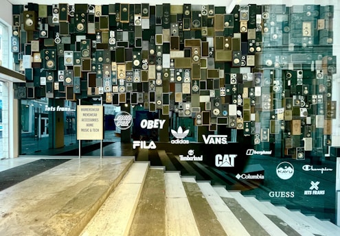 A wall art installation composed of numerous vintage speakers of various sizes and designs, interspersed with light-up brand logos such as Adidas, Vans, and Timberland, among others. The display includes a sign listing product categories like womenswear, menswear, and accessories. The setting appears to be a modern retail environment, featuring reflective surfaces and glass elements.