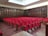 A large room with rows of red cushioned chairs, arranged in an auditorium style facing a wooden podium. The walls are lined with wooden bookshelves filled with numerous books, giving the atmosphere of a library or lecture hall. The floor is covered with wooden flooring, adding warmth to the setting.