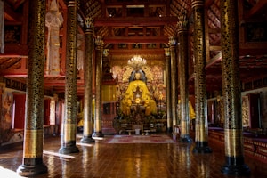A richly decorated temple interior featuring a large, central Buddha statue surrounded by ornate pillars and intricate carvings. The temple is adorned with gold and red colors, and a chandelier hangs from the ceiling. The floor is polished, reflecting the light, and the ambiance is serene and reverent.