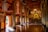 An intricately decorated temple interior with ornate golden columns and detailed wall hangings, featuring a central large golden Buddha statue. The room is illuminated with natural light and chandeliers, creating a peaceful ambiance. A statue of a young monk holding a bowl stands near the entrance, adding to the spiritual atmosphere.