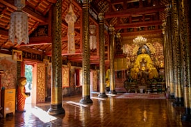 An intricately decorated temple interior with ornate golden columns and detailed wall hangings, featuring a central large golden Buddha statue. The room is illuminated with natural light and chandeliers, creating a peaceful ambiance. A statue of a young monk holding a bowl stands near the entrance, adding to the spiritual atmosphere.