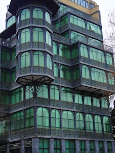 A multi-story building with distinctively green-tinted windows and decorative metal frames. The design features several protruding sections that create a layered effect, with each level having arched windows and intricate latticework. The corner unit is particularly prominent, showcasing a unique architectural style.