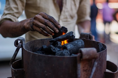 A person is placing pieces of charcoal into a metal container with a small flame visible among the charcoal. The person's hand is smudged with black soot, indicating they have been handling the charcoal. The background is blurred, suggesting a busy environment.