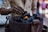 A person is placing pieces of charcoal into a metal container with a small flame visible among the charcoal. The person's hand is smudged with black soot, indicating they have been handling the charcoal. The background is blurred, suggesting a busy environment.