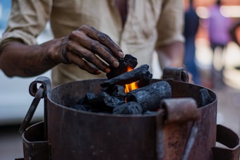A person is placing pieces of charcoal into a metal container with a small flame visible among the charcoal. The person's hand is smudged with black soot, indicating they have been handling the charcoal. The background is blurred, suggesting a busy environment.