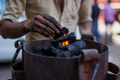 A person is placing pieces of charcoal into a metal container with a small flame visible among the charcoal. The person's hand is smudged with black soot, indicating they have been handling the charcoal. The background is blurred, suggesting a busy environment.