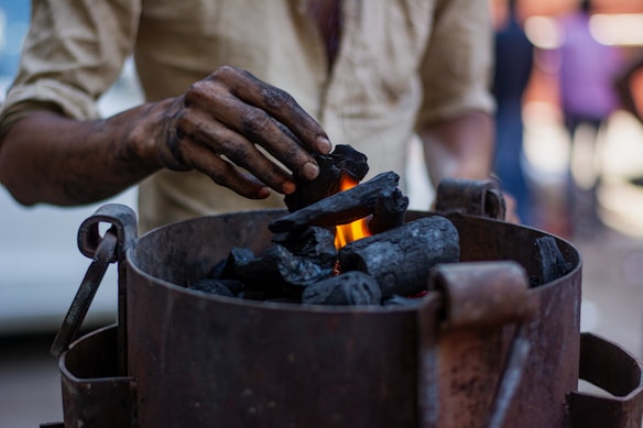 A person is placing pieces of charcoal into a metal container with a small flame visible among the charcoal. The person's hand is smudged with black soot, indicating they have been handling the charcoal. The background is blurred, suggesting a busy environment.