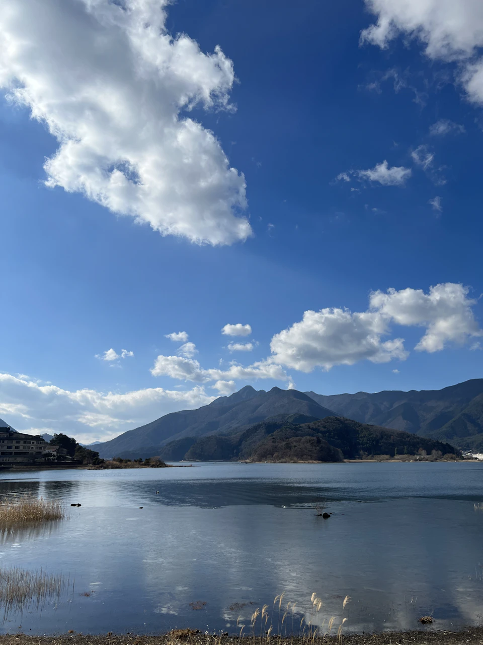 The image depicts a serene lake surrounded by majestic mountains under a clear blue sky with fluffy white clouds. The calm water reflects the beauty of the landscape, and several birds can be seen flying over the lake. In the distance, there are buildings visible on one side of the lake, adding an urban touch to the natural scenery.