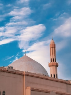 A mosque featuring a large domed roof and a single minaret with a crescent adornment. The building is set against a backdrop of vibrant blue skies with scattered white clouds.