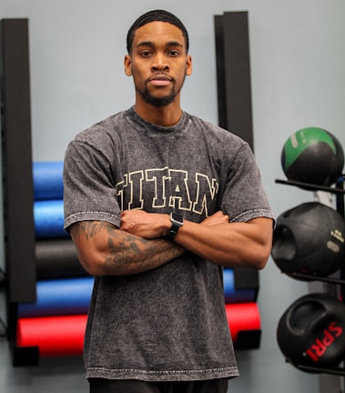 A person stands with arms crossed, wearing a gray t-shirt with the word 'TITAN' in large letters across the chest. They have tattoos on their arms and are wearing a smartwatch. Behind the person, there are various gym equipment including colorful exercise mats and medicine balls.