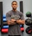 A person stands with arms crossed, wearing a gray t-shirt with the word 'TITAN' in large letters across the chest. They have tattoos on their arms and are wearing a smartwatch. Behind the person, there are various gym equipment including colorful exercise mats and medicine balls.