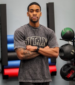 A person stands with arms crossed, wearing a gray t-shirt with the word 'TITAN' in large letters across the chest. They have tattoos on their arms and are wearing a smartwatch. Behind the person, there are various gym equipment including colorful exercise mats and medicine balls.