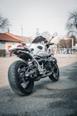 A modern motorcycle is parked on a paved street, with a focus on its rear and side profile. The motorcycle has a sleek design with a white body and dark accents. In the background, there are urban elements like trees, a building, and some utility poles, suggesting an urban or suburban setting.