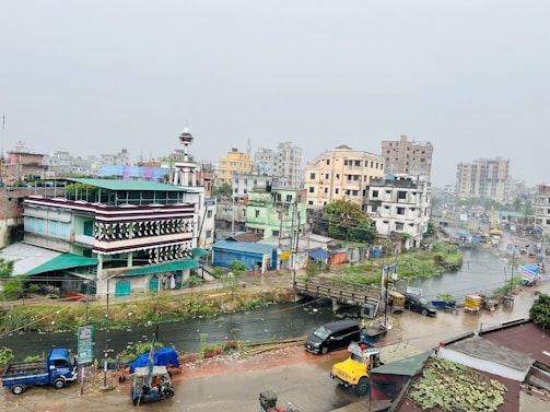 An urban area with a mix of buildings in various states of construction and repair. The foreground features a multi-story building with distinctive architectural elements, including a dome on top. Streets are busy with vehicles including cars, rickshaws, and a yellow truck. There's also visible greenery and a water channel running through the scene. The overcast sky suggests a subdued atmosphere.