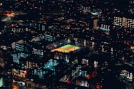 Aerial view of a city at night, showcasing illuminated buildings and streets. A brightly lit sports field is prominently visible in the center, surrounded by apartment complexes and office buildings with lights on.