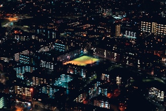 Aerial view of a city at night, showcasing illuminated buildings and streets. A brightly lit sports field is prominently visible in the center, surrounded by apartment complexes and office buildings with lights on.