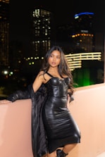 A woman wearing a black leather dress and high heels is leaning against a peach-colored wall. The setting is a nighttime urban environment with blurred city lights in the background. Her long hair flows over her shoulders as she poses confidently.
