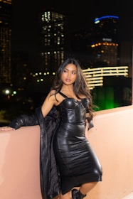 A woman wearing a black leather dress and high heels is leaning against a peach-colored wall. The setting is a nighttime urban environment with blurred city lights in the background. Her long hair flows over her shoulders as she poses confidently.