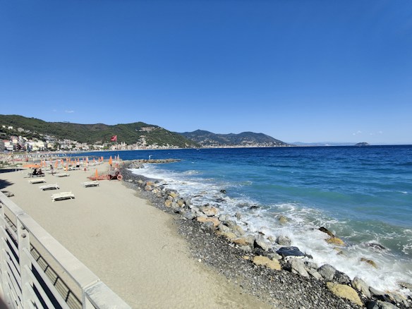A serene beach scene with empty loungers and closed umbrellas under a clear blue sky. Waves gently lap against the rocky shoreline. A small town with colorful buildings lines the coast, backed by lush green hills.