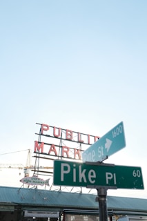 A street sign for Pike Place and Pine Street is prominently displayed in front of a market with the iconic 'Public Market' sign. The sky is clear and blue, creating a backdrop for the metallic structures. A fish market sign is visible beneath the main market sign, indicating a popular urban location.