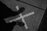 A pair of casual shoes on a person's feet standing on a concrete sidewalk. The ground features a large white 'X' mark, and the photo is captured in black and white, emphasizing texture and shadow.