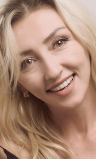 A person with long blonde hair is smiling, showing their teeth. The background is plain, emphasizing the subject's face. The lighting is soft, highlighting the features of the individual.