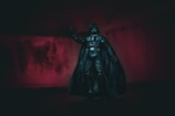 A dark figurine in the shape of a well-known science fiction character stands against a dimly lit background with red tones. The figure is dressed in a black suit with a cape, helmet, and control panel on the chest, embodying a futuristic and villainous appearance.