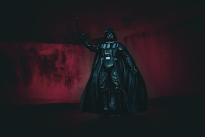 A dark figurine in the shape of a well-known science fiction character stands against a dimly lit background with red tones. The figure is dressed in a black suit with a cape, helmet, and control panel on the chest, embodying a futuristic and villainous appearance.