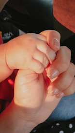 An adult hand gently holding a baby's hand, conveying a sense of care and protection. The nails are neatly manicured with a subtle glitter finish.