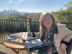 A woman with long blonde hair sits at an outdoor table, writing in a notebook with a pen. She is wearing glasses and a beige jacket over a teal blouse. On the table, there is an open book, a tablet, and a vase with two yellow flowers. The background shows a scenic view of a city skyline under a clear blue sky, with trees and a river in the middle distance.