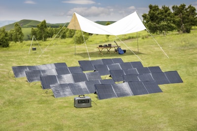 In a grassy outdoor setting, multiple solar panels are arranged on the ground near a white canopy tent. Under the canopy are a table with electronic devices and a blue guitar, suggesting a relaxed, off-grid setup.