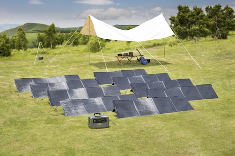 In a grassy outdoor setting, multiple solar panels are arranged on the ground near a white canopy tent. Under the canopy are a table with electronic devices and a blue guitar, suggesting a relaxed, off-grid setup.