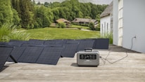 A series of solar panels laid out on a wooden deck are connected to a portable power station. The setup overlooks a scenic landscape featuring lush green fields, trees, and a few rustic houses in the background, suggesting a rural or countryside setting.