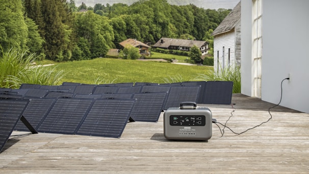 A series of solar panels laid out on a wooden deck are connected to a portable power station. The setup overlooks a scenic landscape featuring lush green fields, trees, and a few rustic houses in the background, suggesting a rural or countryside setting.