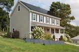 A two-story house with beige siding and a solar panel system on the roof is surrounded by a lush green lawn and various shrubs. There are additional solar panels installed on the ground in front of the house. Large trees are in the background providing a natural setting.