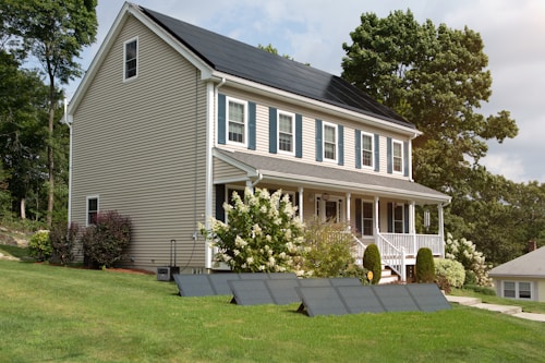 A two-story house with beige siding and a solar panel system on the roof is surrounded by a lush green lawn and various shrubs. There are additional solar panels installed on the ground in front of the house. Large trees are in the background providing a natural setting.