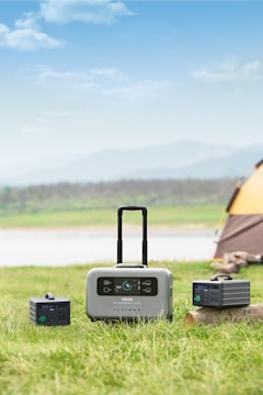 A portable power station with a handle is positioned on a grassy field next to two smaller power units, under a partly cloudy sky. There is a tent in the right background, suggesting a camping setting near a body of water and distant hills.