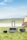 A portable power station with a handle is positioned on a grassy field next to two smaller power units, under a partly cloudy sky. There is a tent in the right background, suggesting a camping setting near a body of water and distant hills.