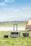 A portable power station with a handle is positioned on a grassy field next to two smaller power units, under a partly cloudy sky. There is a tent in the right background, suggesting a camping setting near a body of water and distant hills.