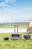 A portable power station with a handle is positioned on a grassy field next to two smaller power units, under a partly cloudy sky. There is a tent in the right background, suggesting a camping setting near a body of water and distant hills.