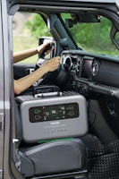 A portable power station is placed on the passenger seat of a vehicle, next to a driver who is holding the steering wheel. The interior of the car features modern design elements with a touch screen display on the dashboard and air vents. The background outside the car appears blurred and green, suggesting a natural environment.