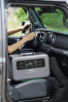 A portable power station is placed on the passenger seat of a vehicle, next to a driver who is holding the steering wheel. The interior of the car features modern design elements with a touch screen display on the dashboard and air vents. The background outside the car appears blurred and green, suggesting a natural environment.