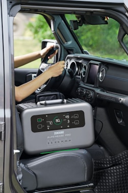 A portable power station is placed on the passenger seat of a vehicle, next to a driver who is holding the steering wheel. The interior of the car features modern design elements with a touch screen display on the dashboard and air vents. The background outside the car appears blurred and green, suggesting a natural environment.
