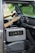 A portable power station is placed on the passenger seat of a vehicle, next to a driver who is holding the steering wheel. The interior of the car features modern design elements with a touch screen display on the dashboard and air vents. The background outside the car appears blurred and green, suggesting a natural environment.