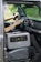 A portable power station is placed on the passenger seat of a vehicle, next to a driver who is holding the steering wheel. The interior of the car features modern design elements with a touch screen display on the dashboard and air vents. The background outside the car appears blurred and green, suggesting a natural environment.