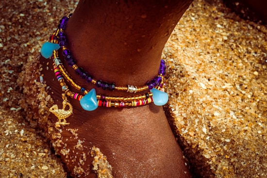 A close-up of an ankle adorned with colorful beaded anklets, featuring blue and purple beads, gold accents, and a small gold charm in the shape of a bird. The foot is partially buried in coarse, golden sand.