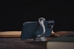 A smartphone is supported by a metallic snake-shaped stand on a wooden surface. The background is dark, and the phone is surrounded by books and papers.