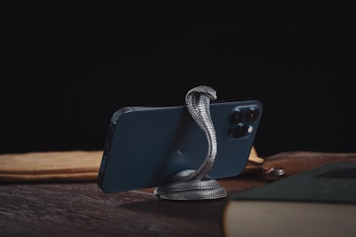 A smartphone is supported by a metallic snake-shaped stand on a wooden surface. The background is dark, and the phone is surrounded by books and papers.